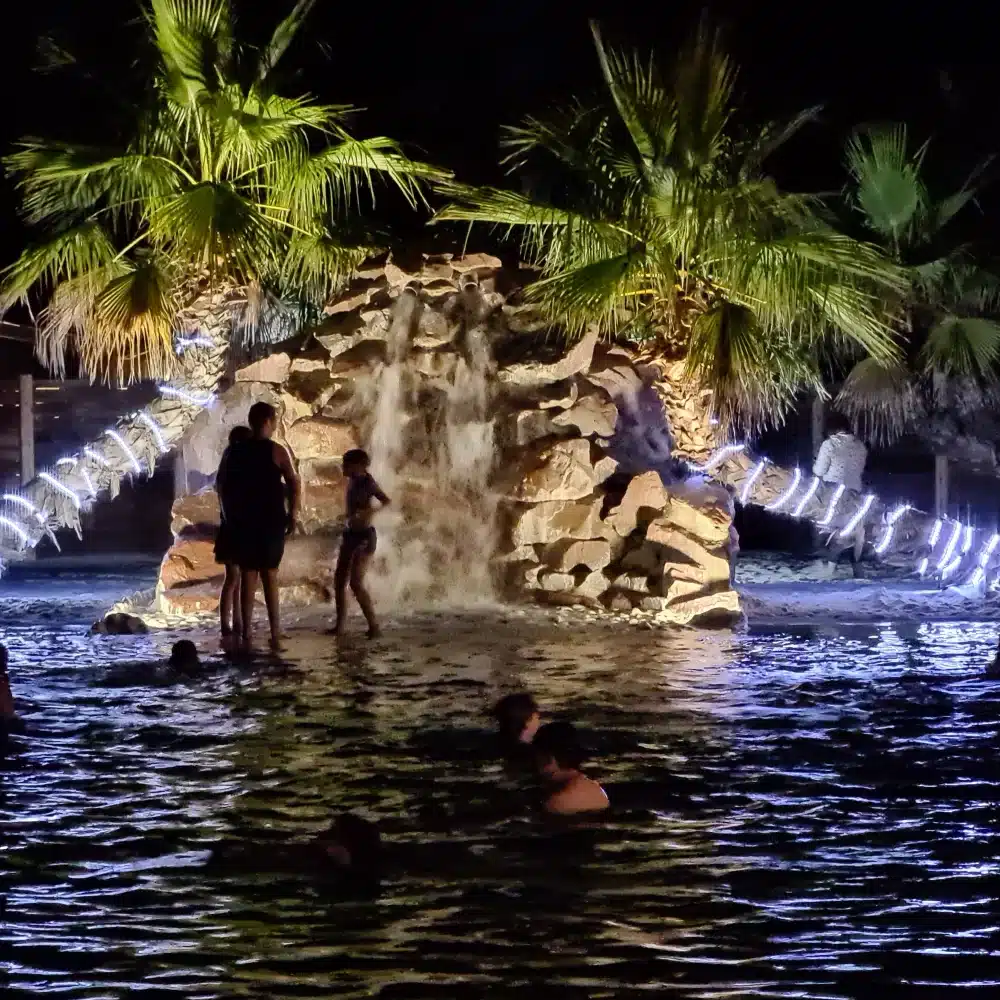 Piscine nocturne avec cascade et palmiers éclairés.
