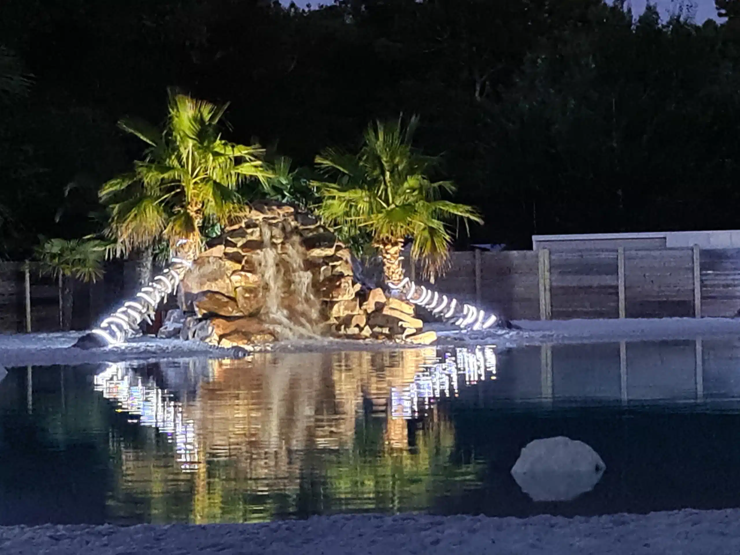 Beleuchteter Wasserfall mit Palmen und Teich bei Nacht.